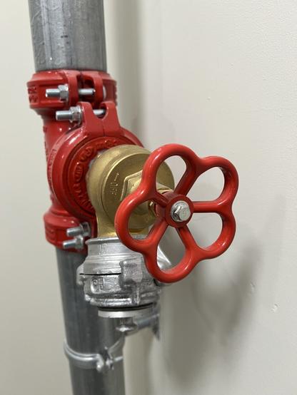 A close-up of a red industrial valve attached to a metallic pipe, featuring a gold-colored lever for operation. The background is a plain wall, emphasizing the valve's functionality.