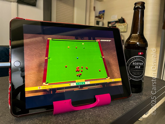 Ein Tablet-Computer zeigt ein Snooker-Spiel auf einem grünen Billardtisch. Das Spiel scheint live zu sein, und der Punktestand ist sichtbar. Rechts neben dem Tablet steht eine braune Flasche Scotch Ale. Das Tablet ist in einem pinken Ständer. Im Hintergrund ist ein unscharfer Blick auf einen Wohnraum zu sehen.