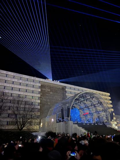 Zu sehen ist der Karl-Marx-Kopf in eine Bühne integriert. Auf dem Dach des Gebäudes dahinter ist ein Teil eines Orchesters mit Blasinstrumenten zu sehen, sowie Teile einer Lasershow.