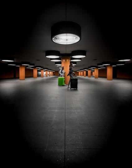 The image shows an underground passage with a symmetrical arrangement of orange pillars supporting the ceiling. The ceiling features round black lights that emit bright white light and are evenly spaced. The floor is covered with dark gray, matte tiles. In the foreground, two individuals walk with rolling suitcases, one of which is a bright green, highly noticeable piece of luggage. The perspective is central, enhancing the depth of the space as the pillars and lights repeat into the distance. The scene appears calm and organized, with no visible crowds.