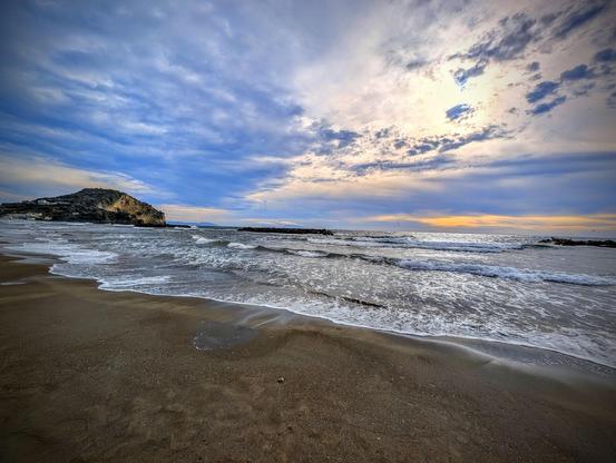 The image depicts a coastal scene with a sandy beach in the foreground and waves gently lapping at the shore. In the background, there are rocky outcroppings or possibly small islands. The sky is mostly cloudy with patches of blue and a hint of orange or yellow, suggesting the time might be around sunrise or sunset. The ocean appears calm, and the overall atmosphere is serene and picturesque.