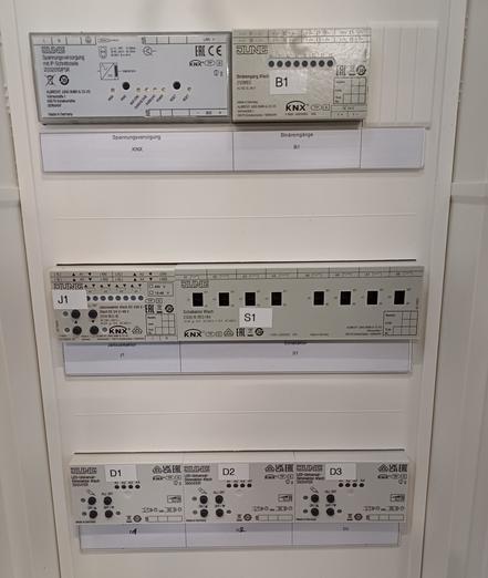 A control cabinet with several installed KNX components. The modules are labeled and arranged in rows, including power supply, binary inputs, switch actuators, and dimmer modules.