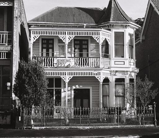 Monochrome. A two storey 19th century white weatherboard house that has seen better days. It has balconies on both levels with elegant and elaborate railings in wooden fretwork. On the upper railing is a largish handwritten sign on what looks like a sheet which says “Sinking Ship”