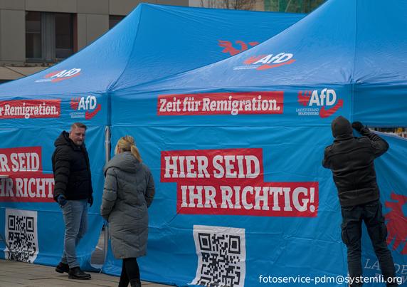 drei personen bestücken ein pavillion mit seitenfolie mir afd werbung: "zeit für remigration - hier seid ihr richtig - afd"