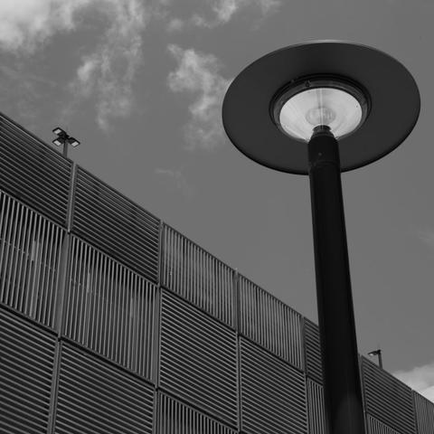 Black and white photograph looking up at a multi-storey car park.  The side of the carpark is made of square patches of steel strips. Each patch has strips running vertically or horizontally, making a chequerboard pattern on the side of the building.  A light pole in the foreground peaks above the car park behind.