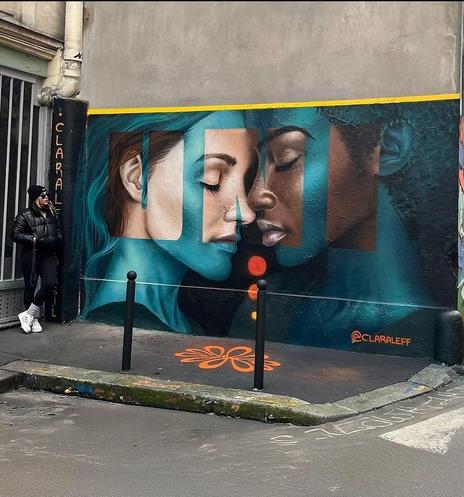 Streetartwall. A beautiful mural with two women's heads was sprayed/painted on a street wall in Paris. The realistically designed mural shows a light-skinned young woman with dark hair standing opposite a dark-skinned young woman. Both have their eyes closed and their noses are slightly touching. The picture is transparently covered with green paint. The green is only omitted in the form of the word "Uni" in capital letters, revealing the view of the two of them. The artist, a young blonde woman in winter clothing, cap and sunglasses, stands next to her mural and looks at it. (In front of the mural is a sidewalk with a drawn stylized flower and some iron pillars)