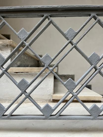 A set of white marble steps, very plain, and their railings. The railings are painted very pale grey. Apart from the top and bottom rail, which follow the line of the stairs, they are widely spaced diagonal bars, single in one direction, double in the other. They cross and form diamonds the size of my hand. At the bottom corner of each diamond there is a smaller diamond of solid metal, about the size of a teabag.