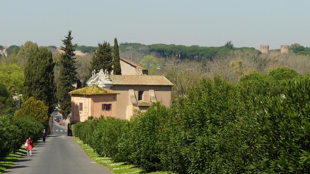 a road runs through a green landscape, past a church. in the distance the two watchtowers of the city gate stand above the trees.