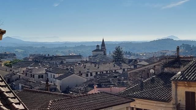 Blick von oben auf die mittelalterliche umbrische Stadt Amelia. Im Vordergrund sieht man die Dächer der Häuser und eine Kirche, im Hintergrund mit Bäumen bewachsene grüne Hügel und einen blauen Himmel.