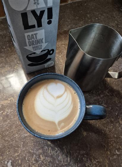 Oat milk latte with latte art surrounded by a carton of oat milk and a milk jug