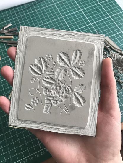 I’m holding a small linoleum plate carved with a character holding a giant strawberry in front of some strawberry plants.