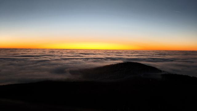 View from the tower on the top of a hill before the sunrise. The sun is just below the horizon creating an orange/yellow horizontal strip. The sky is clear, there is a weather with an inversed temperature, all the dark clouds are below. Small part of the hill is seen as descending down. There view is in the sunrise direction.