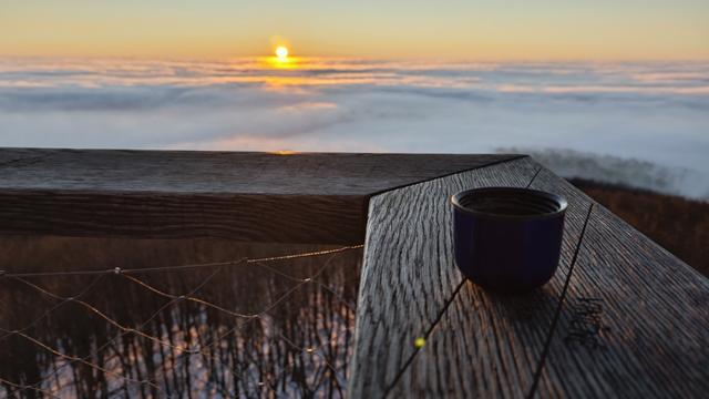 The same place and same viewing direction. Sun is partially above them horizon, there is more light. Part of the wooden fence of the tower (corner) is visible. A small cup is placed on it. Down are trees visible.