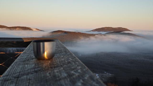 The same place, the viewing direction is opposite, the sun is behind the back. A small stainless steel cup with sun reflection is again placed in the view. The are hills ascending from the fog/clouds lit by the sun.