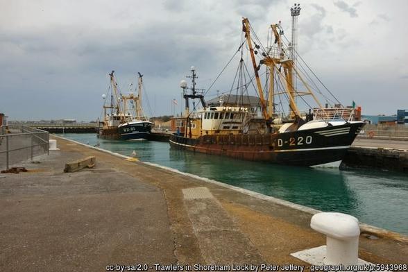 Two steel trawlers in a narrow strip of water between paved areas. A large white-painted bollard in the foreground.