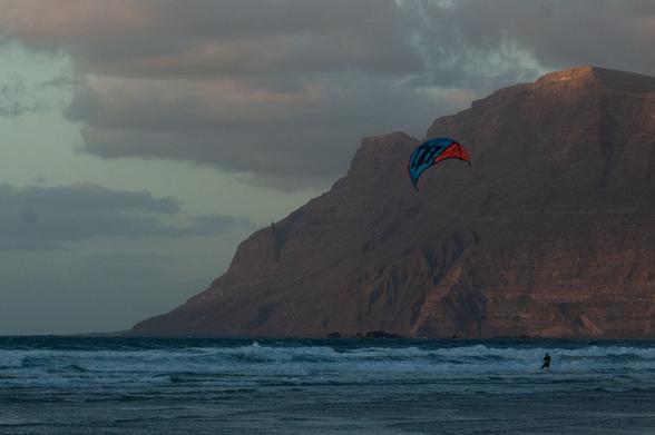 People performing kitesurf with a mountain in the back an a cloudy day in the sunset on the Atlantic Ocean.