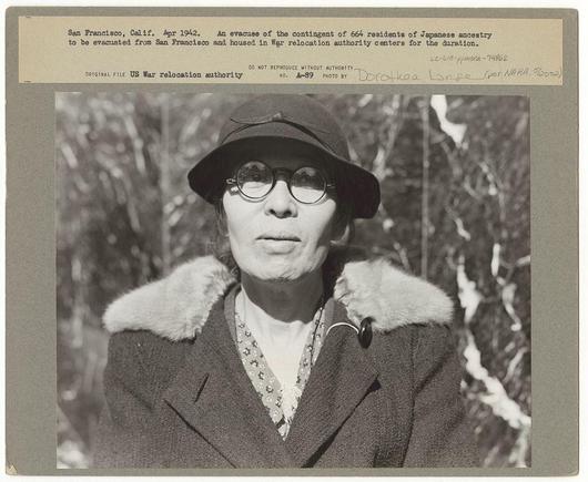 The image is a black-and-white photograph showing an elderly woman with glasses and wearing a dark coat with fur trim. She appears to be outdoors, as indicated by the background foliage visible through gaps in what seems like branches or leaves. The subject wears round-rimmed eyeglasses, has short hair parted on her left side, and is looking directly at the camera.

A caption above the photo reads: "San Francisco, Calif., Apr. 1942. An evacuee of the contingent of 664 residents of Japanese ancestry to be evacuated from San Francisco and housed in War Relocation Authority centers for the duration." Below this text is additional information stating that it's an original file by US War relocation authority with a photo number "A-53" and signed by Dorothea Lange, known as 'Lange' (likely referring to her full name: Dorthea Emma Lang).

The photograph has several pieces of tape indicating its archival nature. A label affixed at the top right corner mentions details about evacuees from San Francisco during World War II, referencing their displacement and internment in war relocation centers.

There is a handwritten note on the bottom left side that reads: "San Francisco - Apr '42 6/17 3-5pm" which could indicate a date or time reference related to this photograph.