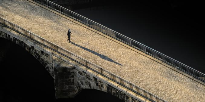 Fotografie von schräg oben auf eine mit Kopfsteinpflaster belegte Brücke, die schräg von oben links nach unten rechts durch das Bild verläuft. Die Brückenpfeiler bestehen aus Sandstein, der teilweise dunkel angelaufen ist. Das Geländer ist aus grauem Metall. Zu den Seiten der Brücke ist dunkelheit. Das Kopfsteinpflaster wird von der niedrig stehenden Sonne golden erleuchtet. Ein dunkel gekleideter Mensch läuft nach rechts über die Brücke, mit einem weißen Smartphone in der Hand. Die Person wirft einen langen Schatten.