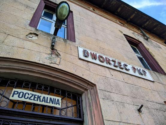 Zbliżenie na fragment niszczejącego budynku. W lewym dolnym rogu górna część drzwi z napisem „Poczekalnia”. Nad drzwiami spora lampa a na prawo od niej napis „Dworzec PKP”.