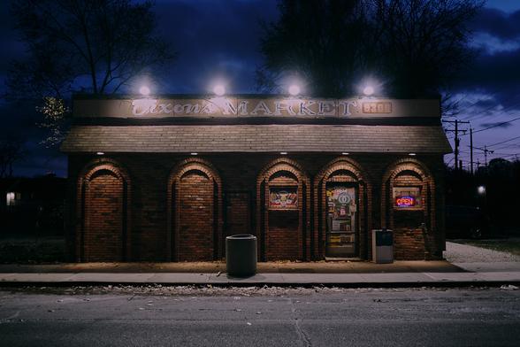 A rectangle building shot shortly after sundown, the background is dark with light fading through the clouds. There are four round bright lights above the store lighting the words "Dixon's Market". The side of the building has five shapes like doors on the side with a rounded top with one of them being the entrance to the building.