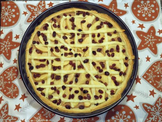 Das Bild zeigt eine runde Apfeltarte in einer Backform. Die Tarte ist mit Apfelscheiben, Rosinen und einem Gitter aus Teigstreifen dekoriert. Sie liegt auf einer weihnachtlich gemusterten Tischdecke mit Sternen- und Schneeflockenmotiven in Rot und Weiß.