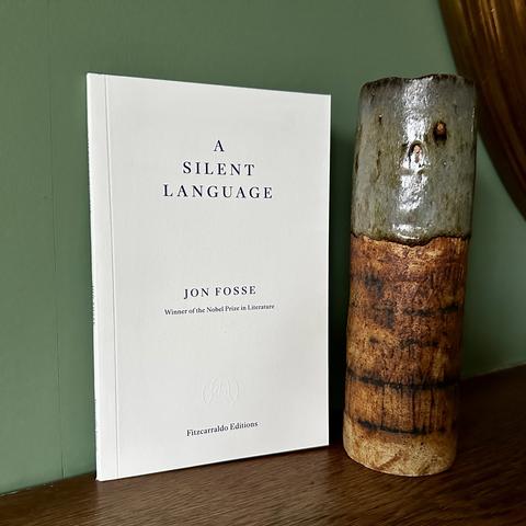 A copy of a slim book, A Silent Language by Jon Fosse. The book stands on a wooden mantlepiece beside a ceramic cylinder in brutalist style, by British potter Alan Wallwork.