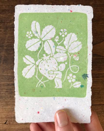Linocut print of a tiny character holding a giant strawberry in front of a few tall strawberry plants. The character is smiling. The picture is printed in bright green ink on handmade light grey paper with a few colored dots in it.