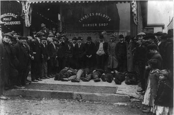 Photograph shows men standing in front of Charles Ohlott's barber shop in Carteret, New Jersey; six wounded men lay on the ground awaiting an ambulance after being injured during a strike at the Williams & Clark Fertilizing Company near Roosevelt, N. J., April 13, 1915. Underwood & Underwood, C. C. (ca. 1915) Strikers at Roosevelt. Wounded awaiting ambulance. New Jersey Carteret, ca. 1915. [Photograph] Retrieved from the Library of Congress, https://www.loc.gov/item/2013647203/.