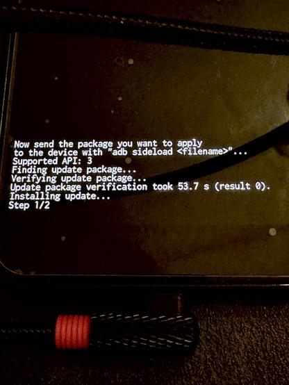 Eine Nahaufnahme eines Handybildschirms, der Befehlszeilen zum Aktualisieren von Software anzeigt. Es werden Meldungen zum Senden eines Pakets, zur Überprüfung des Updates und zur Installation angezeigt, wobei der Fortschritt angezeigt wird. Ein Ladekabel mit einem roten Band ist unten teilweise sichtbar.
