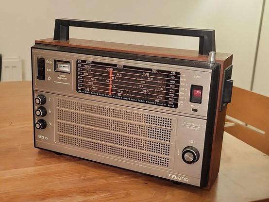 A beautiful 1970s Selena B215 transistor radio with a brushed aluminium effect front panel and wood veneer sides. A sturdy angular carrying handle adorns the top. The front has a long and wide frequency indicator, with markings for five short-wave radio bands, medium wave, long wave and FM. There's an analogue signal strength meter, sturdy knobs for frequency,  volume, bass and treble, chunky black rectangular radio buttons for power, frequency lock and panel lights and a chonker of a rotating dial on the side for band selection.