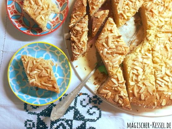 Draufblick auf einen Tisch mit Tischdecke und runden Kuchen auf einer flachen Platte. Der Kuchen ist mit Mandeln verziert und in kleine rautenförmige Stücke geschnitten. Ein Tortenheber liegt teils unter den Stücken. Links sind zwei kleine, gemusterte Tellerchen auf denen jeweils ein Stückchen Kuchen liegt.