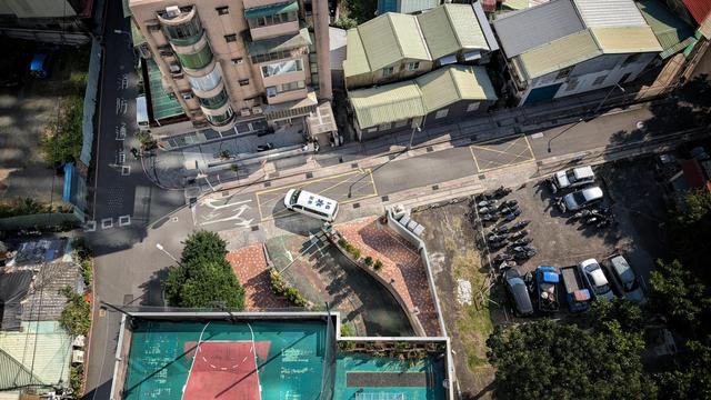 Das Bild zeigt eine städtische Umgebung aus der Vogelperspektive. Es zeigt eine Straßenkreuzung, auf der ein Krankenwagen steht. Links im Bild ist eine Straße mit einem Hinweis auf die Fahrbahn geschrieben, vermutlich "Feuerwehrzufahrt" (消防通道). Rechts neben der Straße gibt es mehrere Gebäude mit unterschiedlichen Dachfarben, überwiegend in Grün und Beige. Auf einem angrenzenden Parkplatz stehen Autos und viele Motorroller.

Im unteren Bereich des Bildes befindet sich ein Basketballplatz mit roten und grünen Spielfeldern, daneben ein kleiner gepflasterter Platz mit Pflanzen. Einige der umliegenden Häuser wirken älter, und die Szenerie ist von Sonnenschein beleuchtet. Die Straßen sind relativ leer, und es sind nur wenige Menschen zu sehen.
