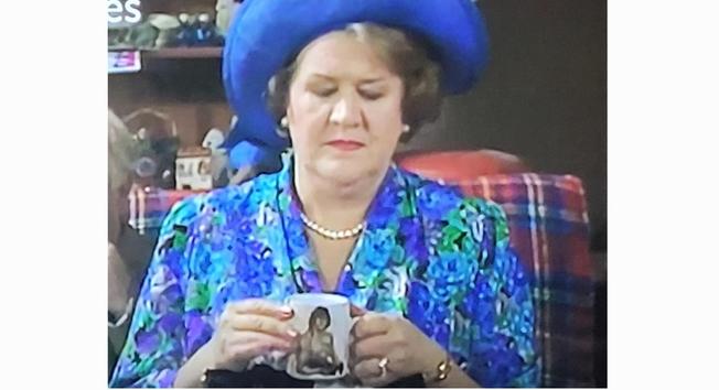 Patricia Routledge as Hyacinth wearing a brightly colored blue flowered dress and matching blue hat looking disdainfuly down at the mug of tea in her hands