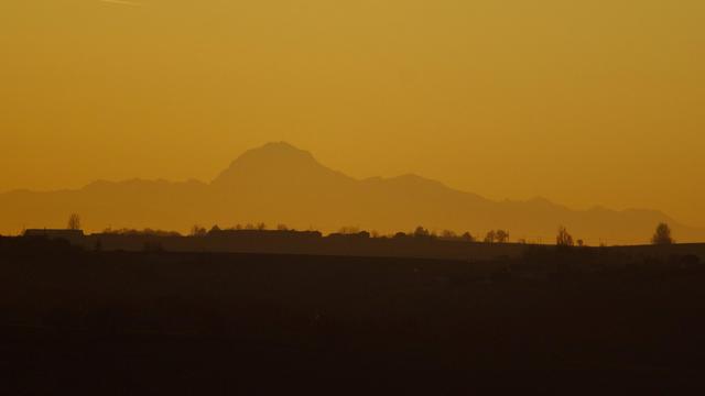 Pic du midi dans un ciel orange pulvérulent, la moitié basse de l'image est constituée de collines dans la pénombre et la ligne d'horizon a des maisons et des arbres. On voit très bien le batiment et l'antenne au somme avec un reflet du soleil sur une vitre surement, qui fait comme une lumiere au sommet.