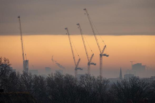 Sunset view of four tall construction cranes, and a fifth further in the distance, behind a line of leafless trees. They stand amid buildings, against a golden sunset sky.