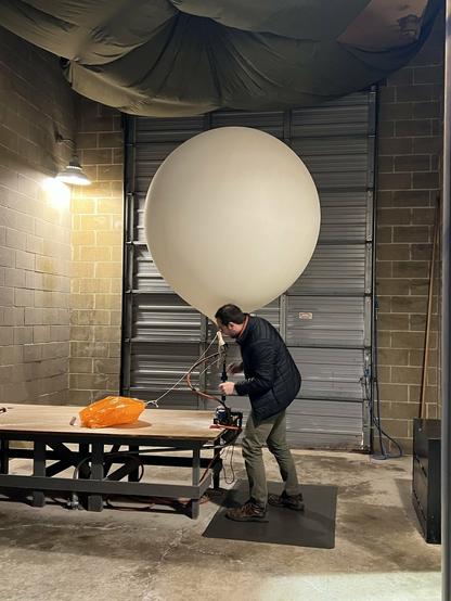 Inside of a garage. An employee of the National Weather Service is preparing a weather balloon for launch. The weather balloon is large (about 6ft in diameter) and white with weather equipment attached to it from below.