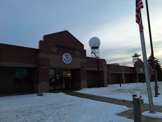 The front of the building for the National Weather Service in Minnesota. A flag pole is in the foreground. Behind the building is a weather radar tower.