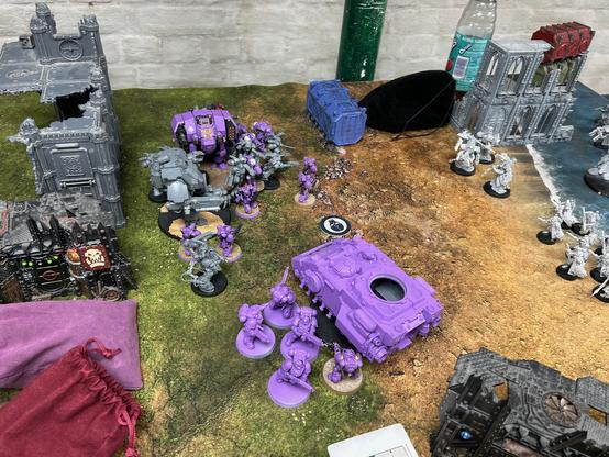 View looking down at my Space Marine army between various bits of terrain and gaming tools. Some of the army are fully painted in purple and gold armour, the rest are in various states from bare grey plastic to mostly finished models. There are two bipedal robots, both have large guns, on has a missile rack and the other has a large hand. A squad of terminators. Two squads of marines with jump packs, a squad of regular marines. A tech marine with various robotic arms on their armour. Five marines in large heavy plate. And a tank with it’s gun turret missing