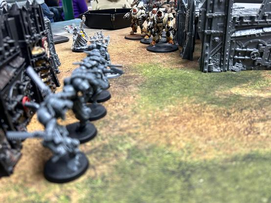Looking between two grey buildings, close to the camera there’s a squad of five unpainted grey space marines with jump-packs and chainswords. In the distance is a squad cream coloured Kastelan Robots and a small squad of white Skitarii.
