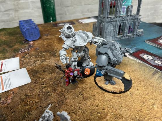 Two large bipedal robots, an Armiger Warglaive and Redemptor Dreadnought) in combat. They are stood next a red TechMarine holding a power axe and plasma weapon. On the Armiger is a orange die showing four.