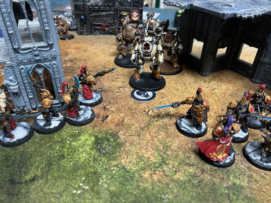 A unit of three cream and one unpainted Kastelan Robots standing on an objective marker between two buildings. Towards the bottom right and left corners are two of my black, gold and red Custodians holding various blue power weapons.