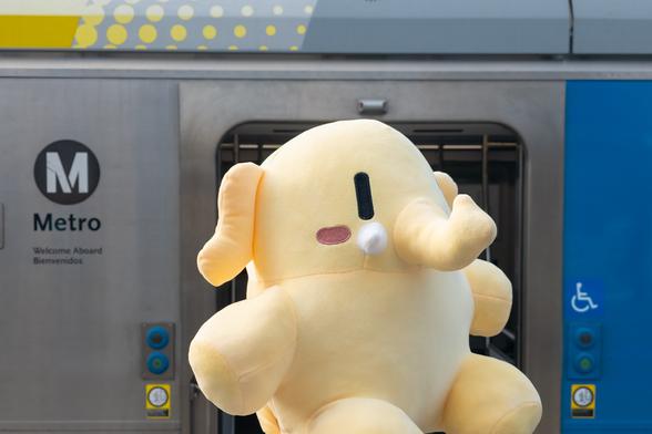 Photo of the Creature Mastodon stuffed toy in front of a Los Angeles Metro light rail train. The train is steel with gray, blue, and yellow paint patterns. The close-up shot of the Creature in the center of the frame largely covers the train's open doors behind it. Towards image-left is the Los Angeles MTA logo and the text, “Metro”, “Welcome Aboard”, “Bienvenidos”.