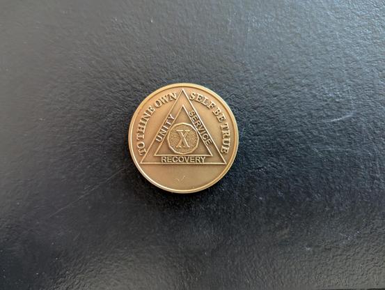 A golden token that has a triangle and circle emblem with a Roman X and the words Unity Service Recovery in the triangle arms. The top two reliefs have the words To Thine Own Self Be True.