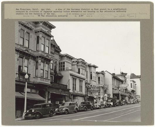 The image is a black and white photograph depicting an urban street scene from San Francisco, California in April 1942. The setting appears to be the historic business district on Post Street within a neighborhood that was previously inhabited by Japanese Americans before their evacuation and housing in War Relocation Authority centers during World War II.

Several old-fashioned buildings with ornate architectural details line both sides of the street. Storefronts, including Nippon Ora Co., Chop Suey, Ching's Drug Company, and Cherryland Hardware store, are visible along the storefront windows. The businesses have signs in English alongside Japanese language text, indicating their diverse customer base.

The streetscape features a row of parked cars from an earlier era, suggesting that this photo was taken during or shortly after World War II when such vehicles were common on American roads. There's also a streetcar visible further down the road and pedestrians walking along the sidewalks.

Above the photograph is handwritten text providing context for its historical significance: "San Francisco, Calif. Apr 1942," followed by additional notes about Japanese ancestry residents before evacuation and housing in war relocation authority centers during World War II. The handwriting indicates that it was possibly recorded or transcribed by someone who had seen this photo at a later date.

The photograph appears to be an origin [...]