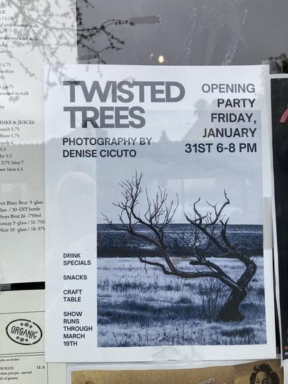 [Image ID: A window with flyers and menus in it. The focus is on a flyer that has a black and white photo of a burnt tree in a meadow of dried grass. The text reads "Twisted Trees photography by Denise Cicuto. Opening Party Friday January 31st 6-8pm. Drink specials, Snacks, Craft table, Show runs through March 19th." End description.]