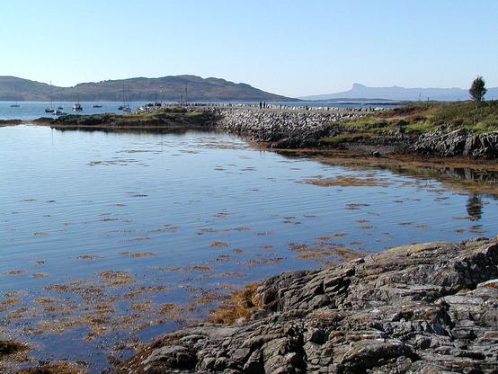 The beautiful view west along Loch nan Ceall from Arisaig near Mallaig to the distinctive outline of the distant island of Eigg. A rocky foreground leads into still waters with floating seaweed, beyond which is a stone pier. There are some moored boats beyond it, with islands in the distance, The sky is blue.