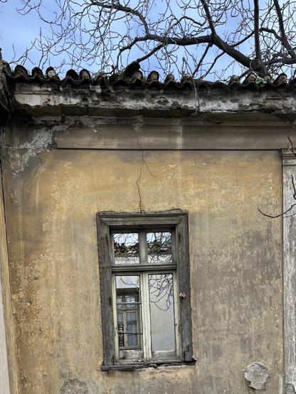 High up on the facade of a hollow shell of an abandoned neoclassical house, an open window. Through it the shuttered window on the opposite wall can be seen, tucked away in the left corner. There’s a strip of sky above the tiled roofline of the house in the image, and another can me seen above the tiled roofline on the other side, through the window