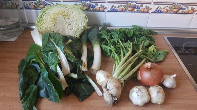 Contenido de la cesta de la semana sobre una encimera de cocina.
Al fondo, medio repollo. Delante, acelgas, puerros, un nabo que parece que sean tres juntos, una cebolla y tres cabezas de ajos.