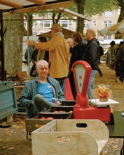 Man seated at a market stall, surrounded by colorful produce and a classic red scale, evoking a warm, lively market vibe. Taken with a Fuji X-T1.