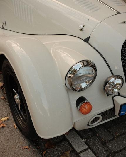 Close-up of a vintage car’s front, focusing on its smooth curves and chrome details, reflecting the surrounding street. Shot with a Fuji X-T1.